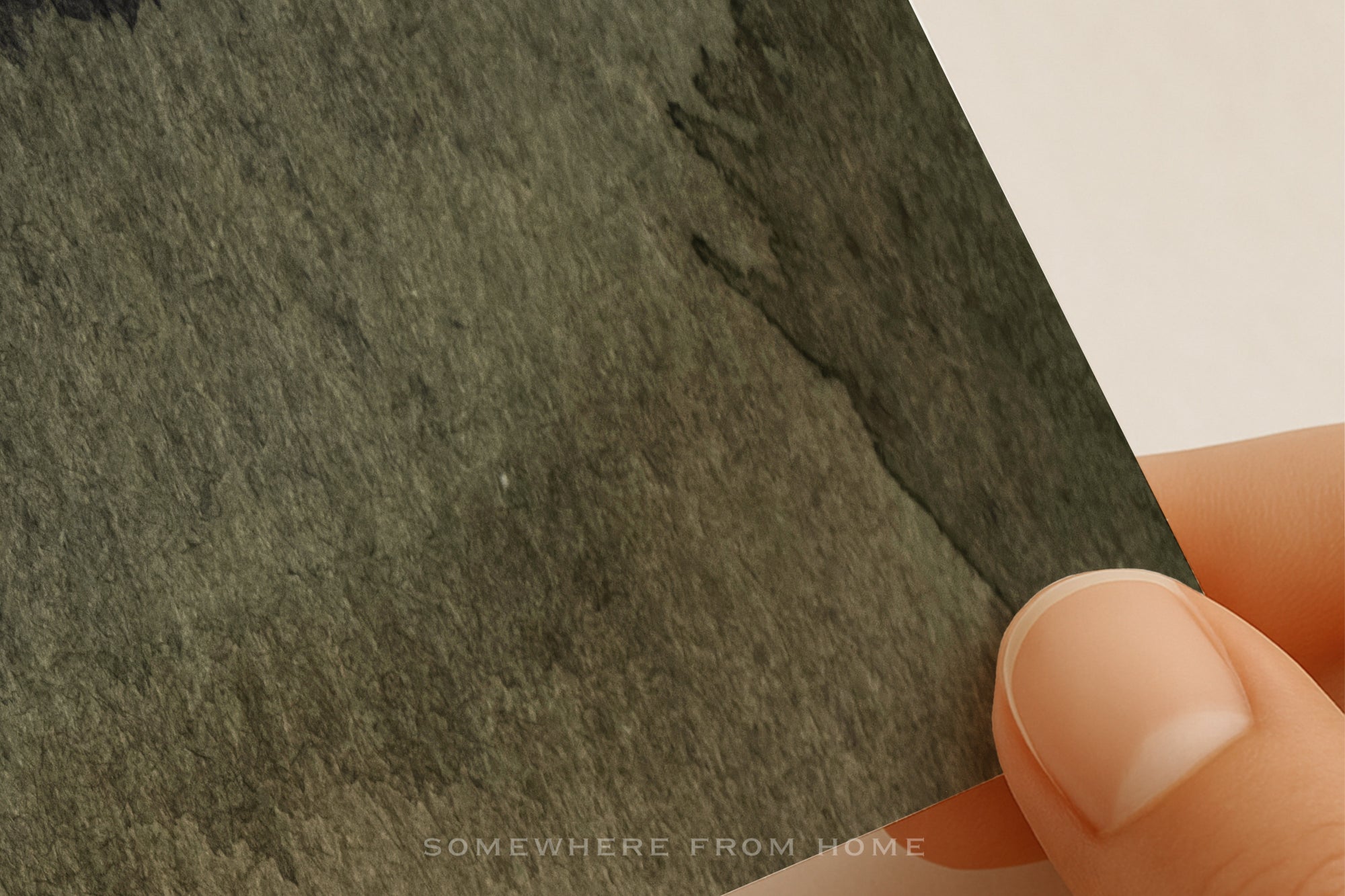 A close-up of a hand holding a textured dark moody olive green abstract watercolor art print 
against a plain white background.