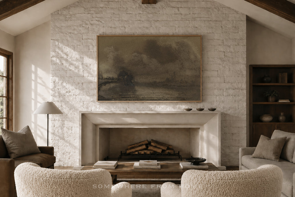A large abstract landscape painting in earthy brown and beige tones hangs on a white brick fireplace wall in a cozy, sunlit living room with textured armchairs and a coffee table.