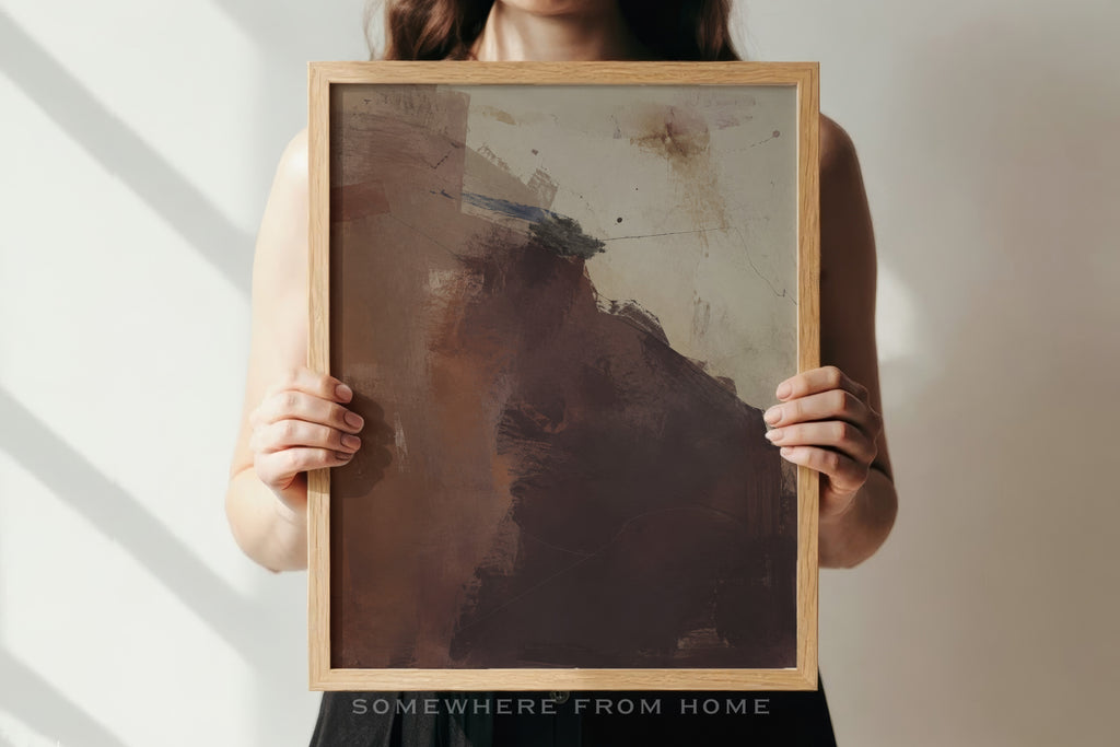 A person holds a framed abstract art print featuring earthy brown, beige, and gray tones. The person is cropped from the shoulders down, holding the light wood frame against a neutral wall with soft diagonal shadows.