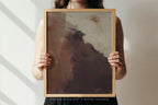 A person holds a framed abstract art print featuring earthy brown, beige, and gray tones. The person is cropped from the shoulders down, holding the light wood frame against a neutral wall with soft diagonal shadows.
