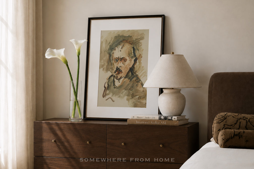 A framed art print featuring a portrait sketch in earthy tones, styled on a dark wood nightstand in a bedroom setting alongside a lamp, a vase of calla lilies, and a bed.