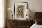 A framed art print featuring a portrait sketch in earthy tones, styled on a dark wood nightstand in a bedroom setting alongside a lamp, a vase of calla lilies, and a bed.