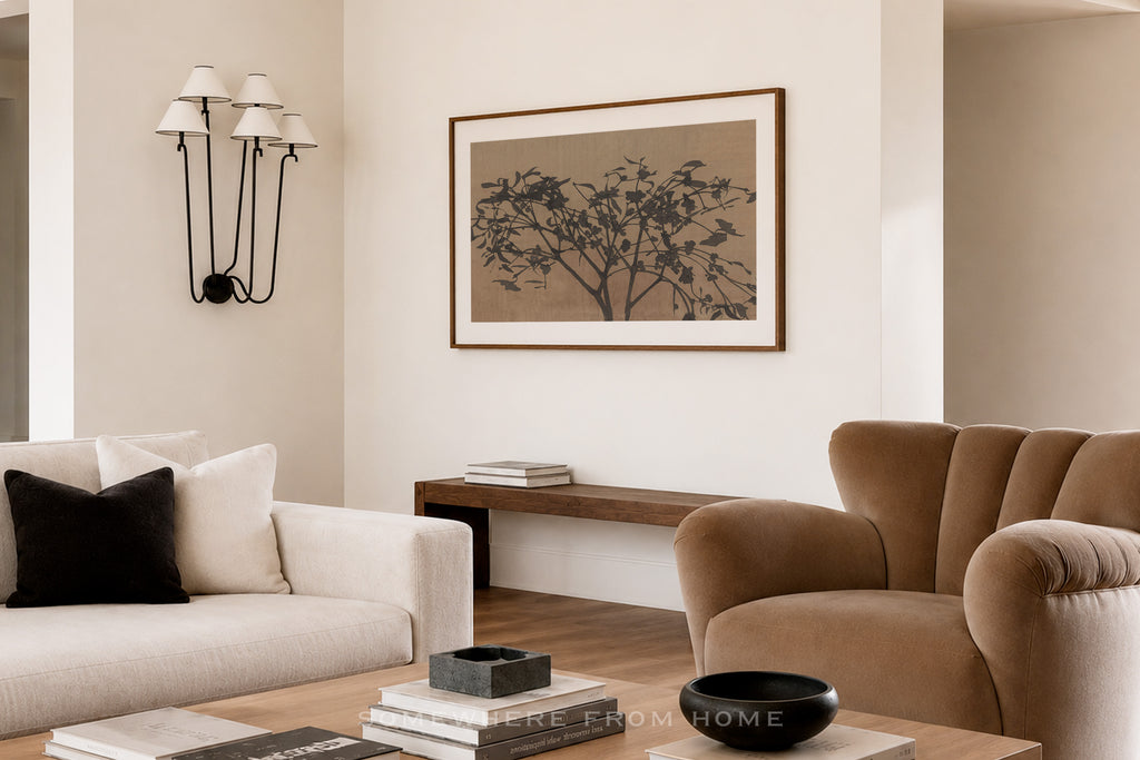 A framed wall art print featuring a dark tree silhouette on a warm beige background, styled in a modern living room setting with a cream sofa, a brown armchair, and a wooden console table.