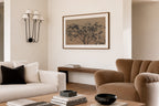 A framed wall art print featuring a dark tree silhouette on a warm beige background, styled in a modern living room setting with a cream sofa, a brown armchair, and a wooden console table.