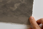 A close-up of a hand holding the edge of a textured mountain landscape art print with a hand torn, rustic border against a plain white background. 
