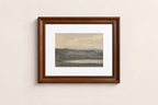 A framed landscape art print with a white mat and a dark brown wooden frame, hanging on a light beige wall.