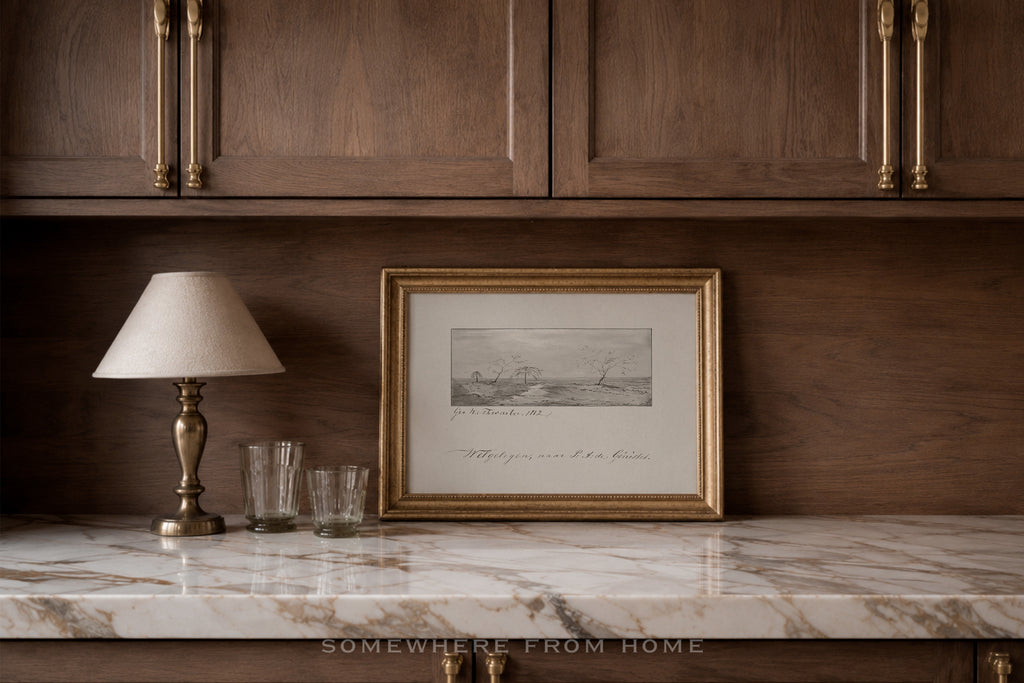 A framed black and white landscape sketch with elegant cursive writing, set in a gold frame on a white marble countertop against a dark wood cabinet background.