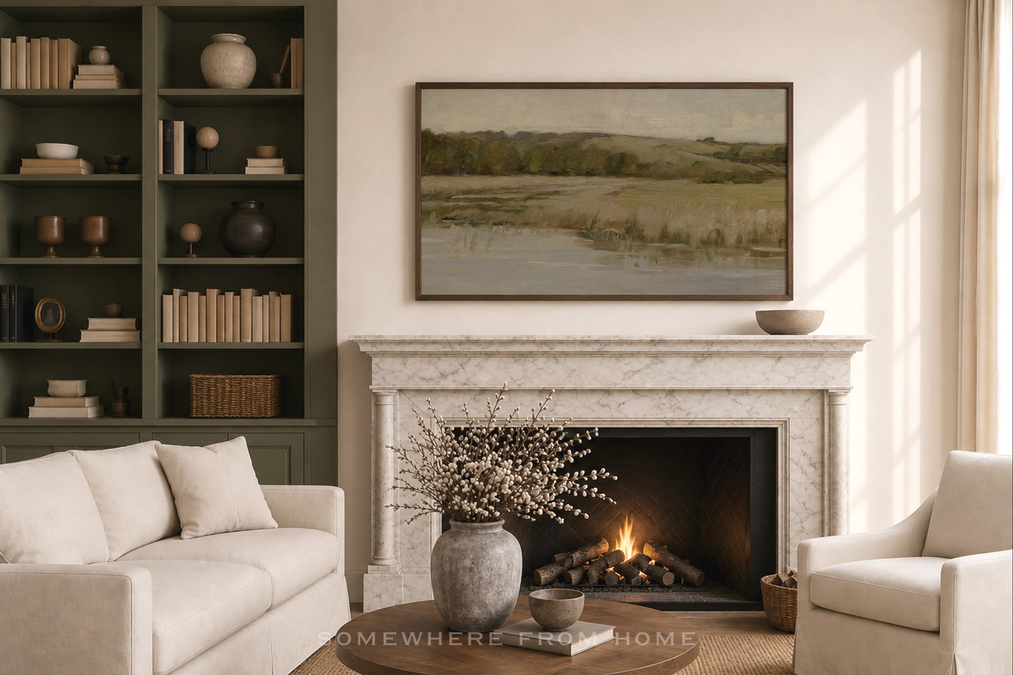 A framed landscape painting hangs above a white marble fireplace in a styled living room with a cream sofa, a matching armchair, a dark green bookshelf, and a lit fire in the fireplace.