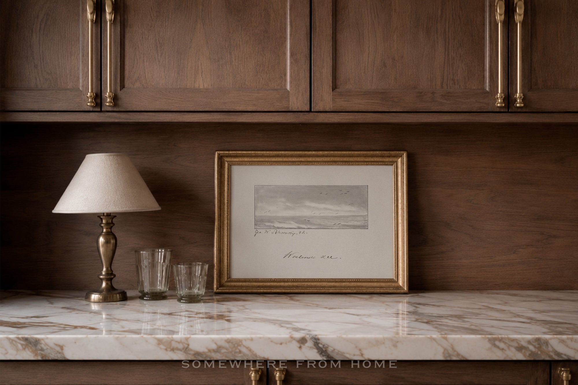 A framed black and white landscape sketch with a gold frame and white mat, displayed on a white marble countertop next to a table lamp and two glasses, set against a dark wood cabinet background.
