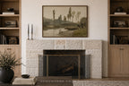 A framed landscape art print hangs above a stone fireplace in a styled room with wooden shelves, candles, books, and a vase.