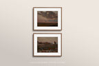 Two framed landscape art prints with white mats and thin brown frames, displayed vertically on a light beige wall. The prints depict moody, dark stormy skies over coastal or rocky terrain.
