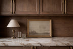 A framed landscape art print with a gold frame sits on a white marble countertop against a dark wood-paneled wall, next to a table lamp and two glasses.