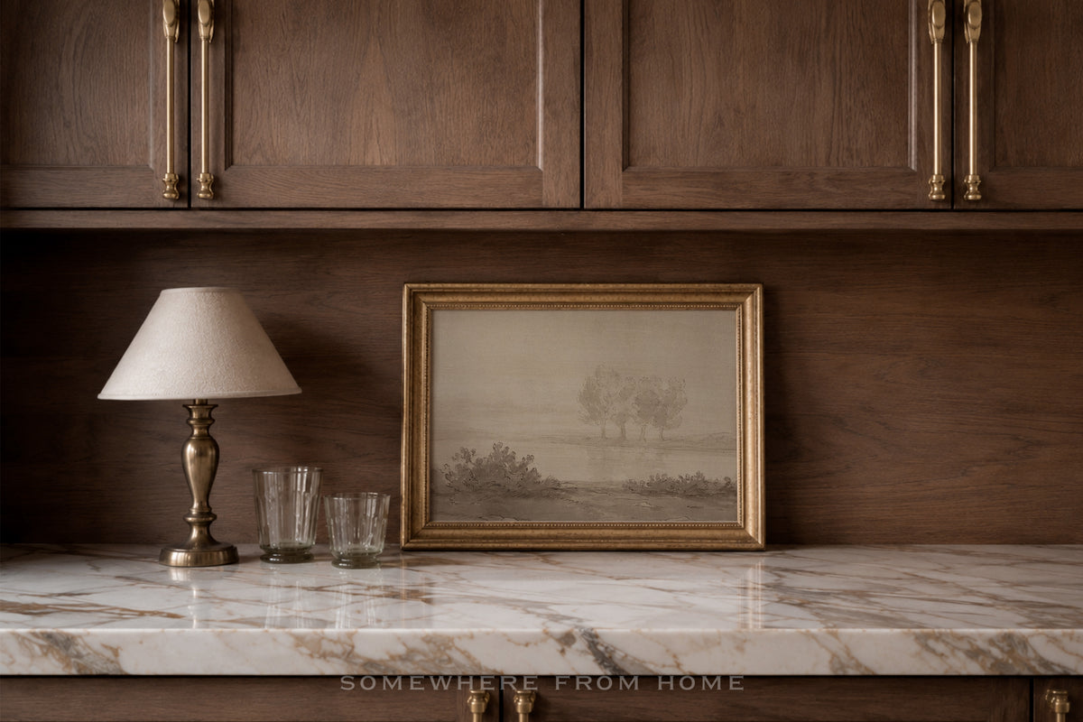 A framed landscape art print with a gold frame sits on a white marble countertop against a dark wood-paneled wall, next to a table lamp and two glasses.