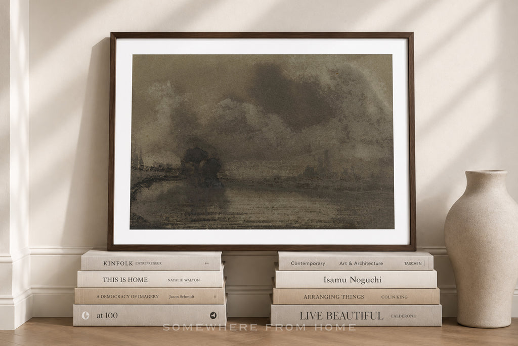 A framed art print featuring a dark, moody landscape painting, styled on a wooden floor with books and a ceramic vase against a beige wall with soft window shadows.