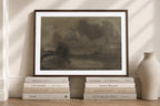A framed art print featuring a dark, moody landscape painting, styled on a wooden floor with books and a ceramic vase against a beige wall with soft window shadows.