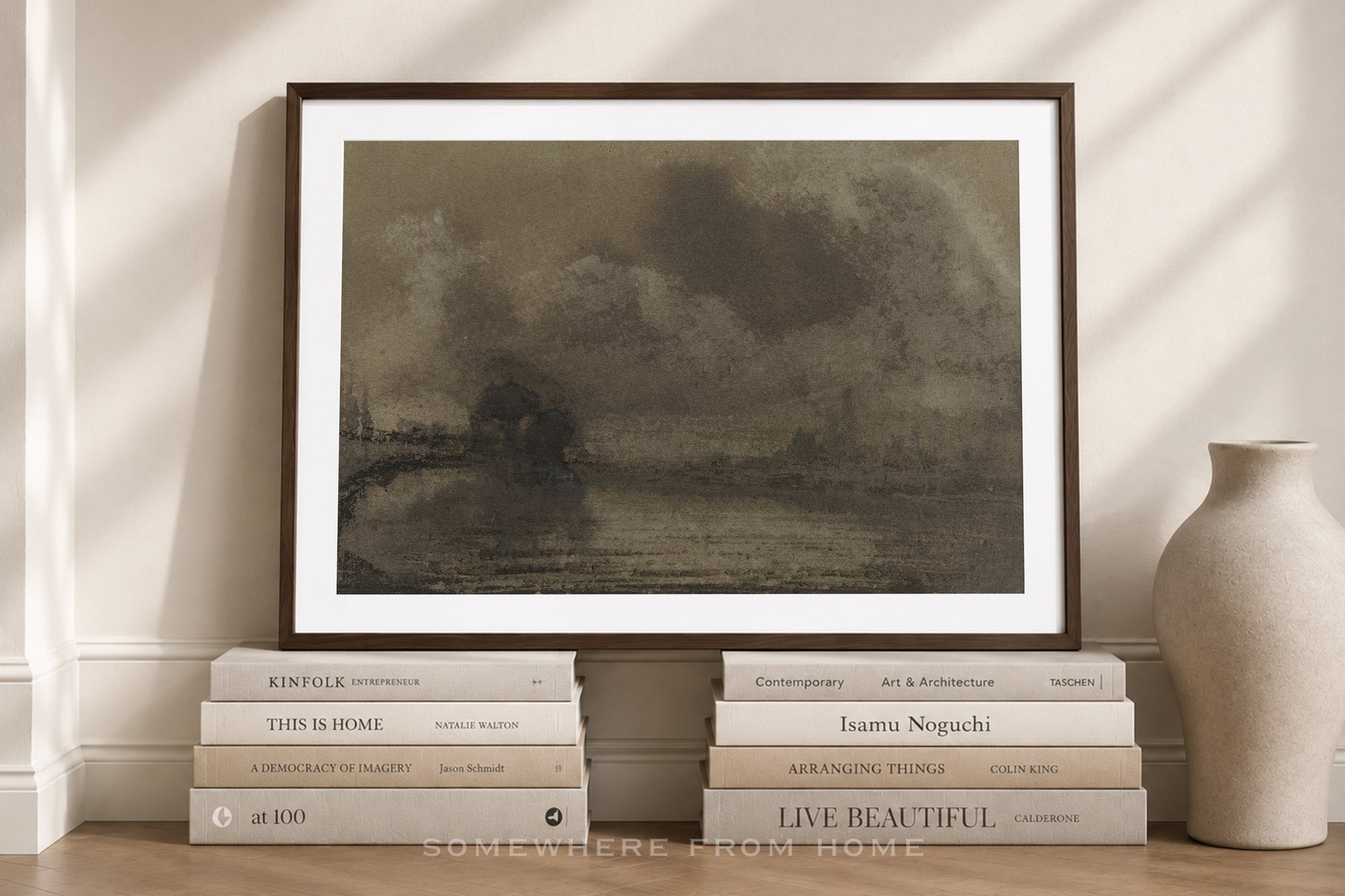 A framed art print featuring a dark, moody landscape painting, styled on a wooden floor with books and a ceramic vase against a beige wall with soft window shadows.