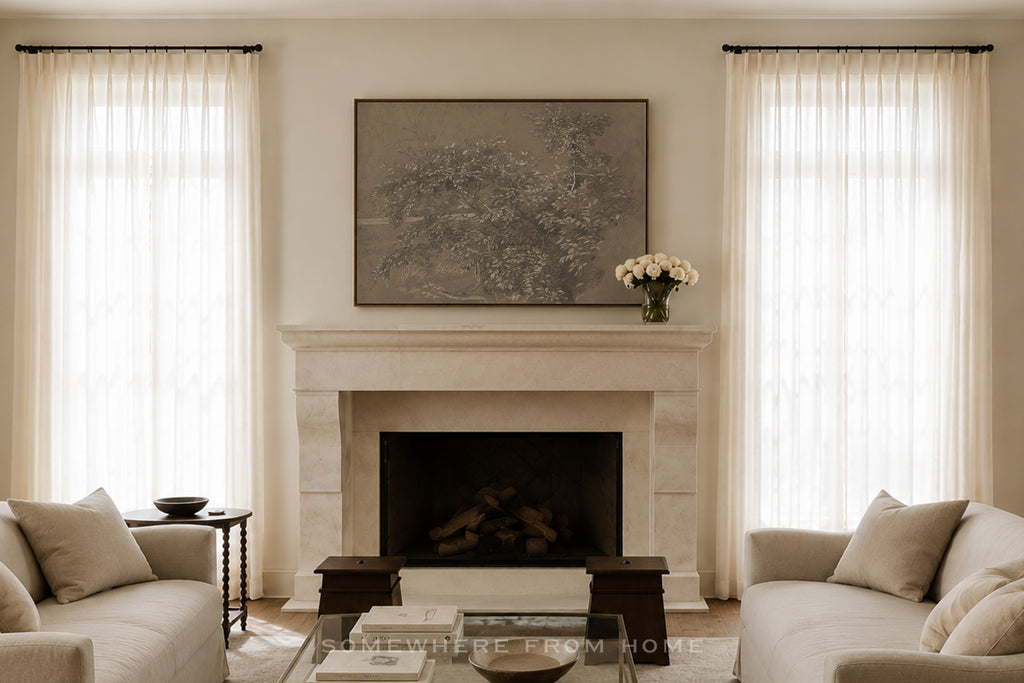 A styled living room featuring a central stone fireplace with a large framed landscape painting above it, flanked by two tall sheer white curtains. Comforter sofas on both sides, a glass coffee table, and a vase of white flowers on the mantel complete the scene.