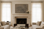 A styled living room featuring a central stone fireplace with a large framed landscape painting above it, flanked by two tall sheer white curtains. Comforter sofas on both sides, a glass coffee table, and a vase of white flowers on the mantel complete the scene.
