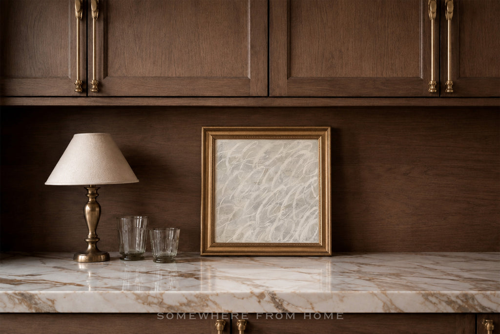 A framed black and white sketch print with a gold frame sits on a white marble countertop against a dark wood cabinet wall, next to a table lamp and two glasses.