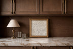 A framed black and white sketch print with a gold frame sits on a white marble countertop against a dark wood cabinet wall, next to a table lamp and two glasses.