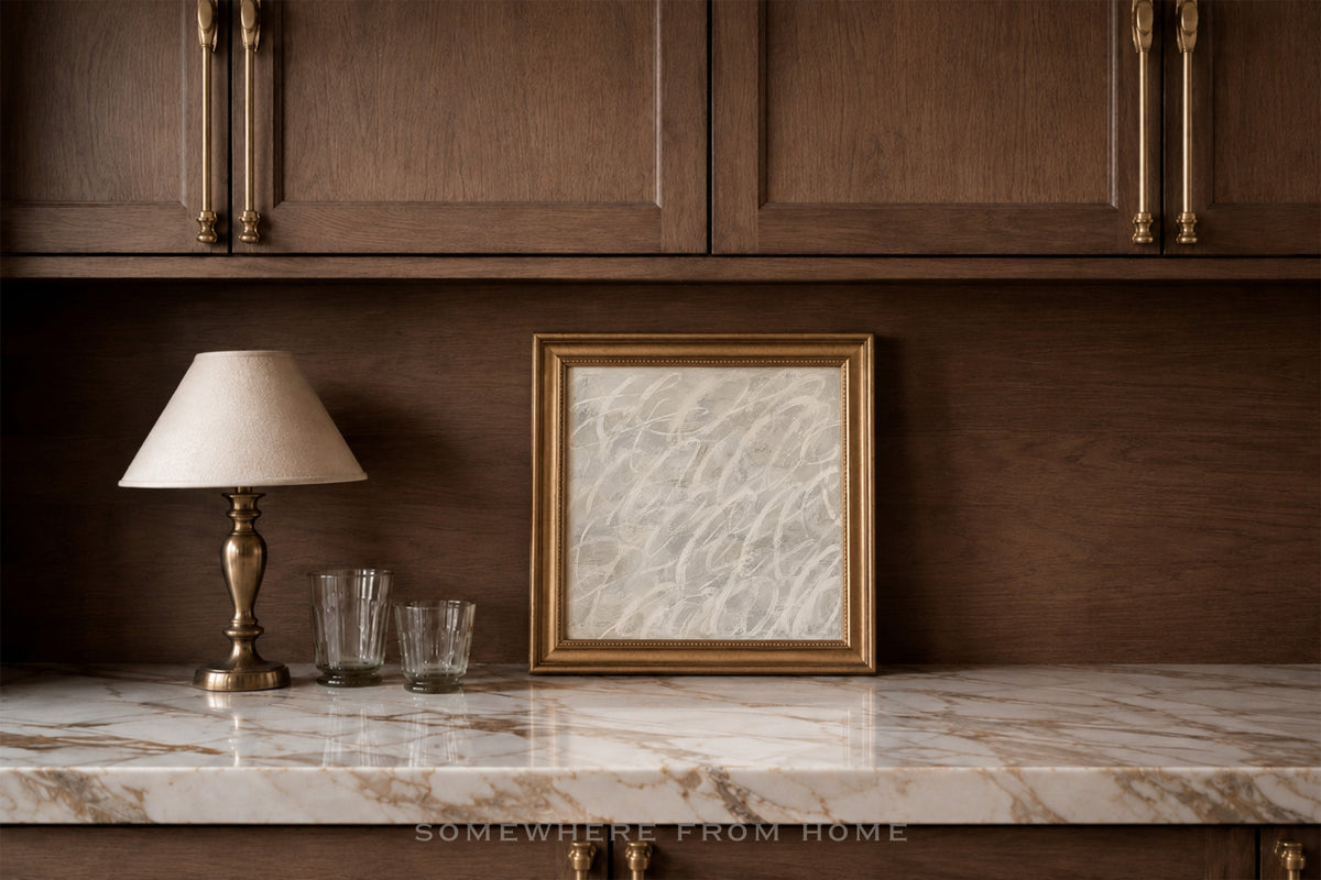 A framed black and white sketch print with a gold frame sits on a white marble countertop against a dark wood cabinet wall, next to a table lamp and two glasses.
