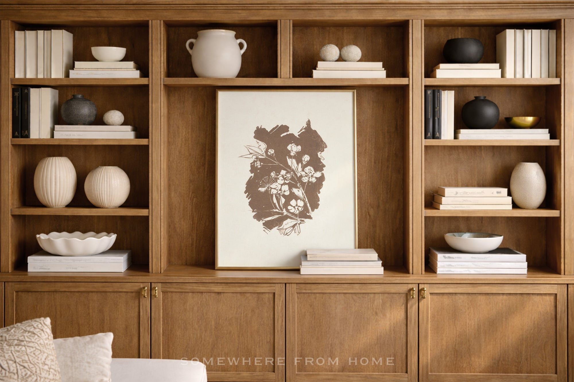 A framed botanical art print with a dark brown floral illustration on an off-white background, displayed on a wooden built-in bookshelf with books and decorative vases.
