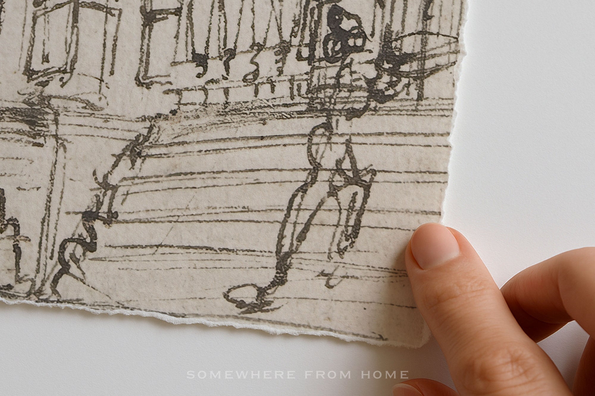 A close-up of a hand holding the edge of a square piece of textured paper with a black ink sketch of a person on a street scene. The paper has a torn edge and is shown against a plain white background.