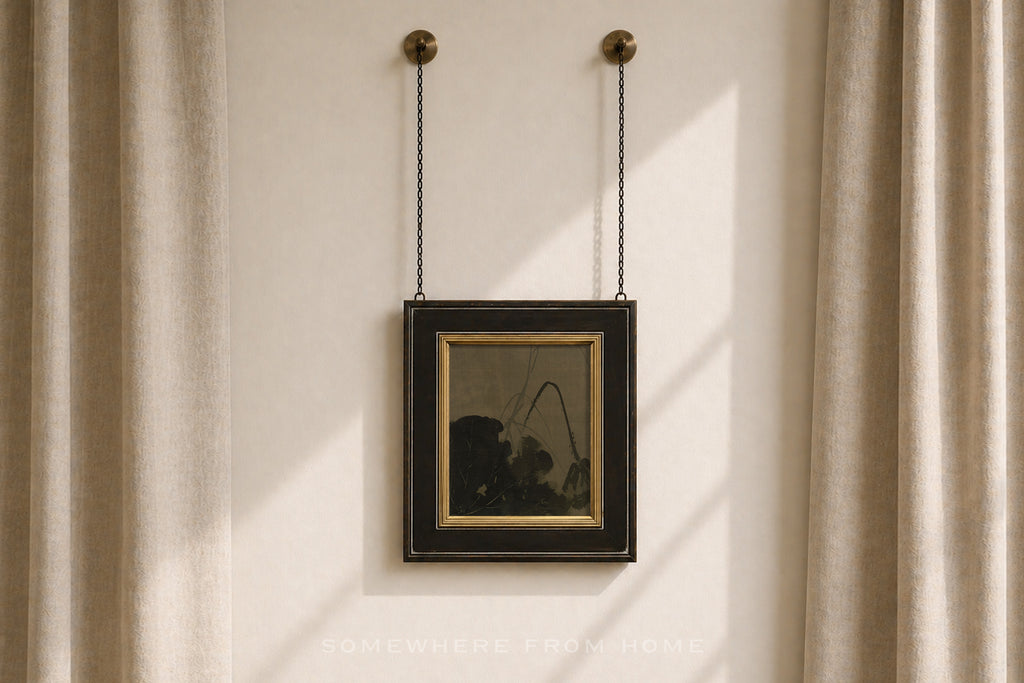 A framed art print with a black frame and gold inner border hangs from two black chains on a beige wall between beige curtains.