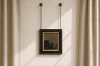 A framed art print with a black frame and gold inner border hangs from two black chains on a beige wall between beige curtains.