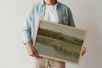 A person holding a framed landscape painting. The painting depicts a muted, pastoral scene with fields and trees in soft beige, green, and gray tones.