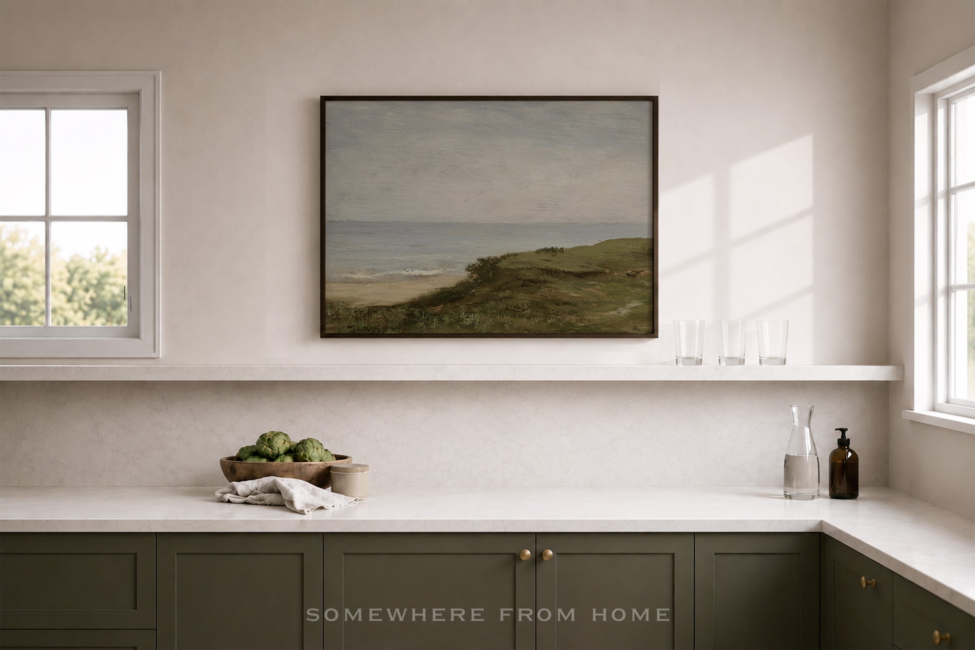 A framed landscape art print depicting green grassy dunes and a distant sea, displayed on a white wall above a kitchen counter in a bright, modern room.