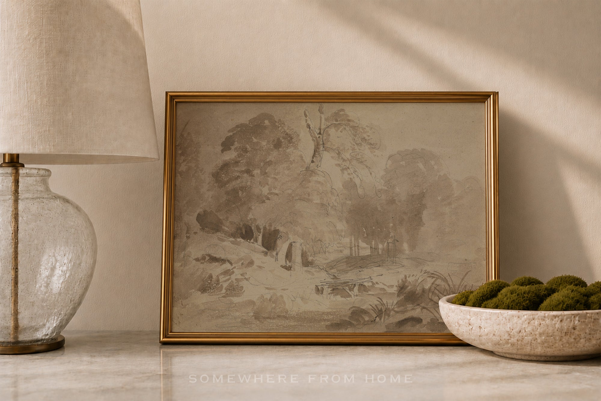 A framed landscape art print with a vintage-style monochrome sketch and a gold frame, styled on a marble surface next to a glass lamp and a bowl of green moss.