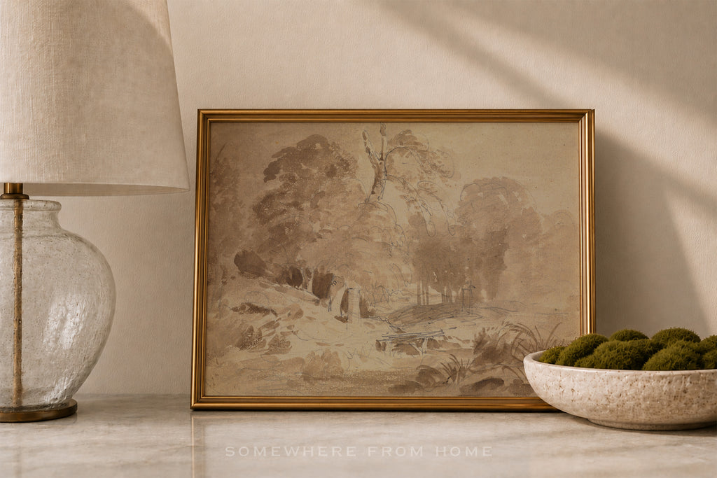 A framed sepia-toned landscape painting with a gold frame, styled on a marble surface next to a glass lamp and a bowl of green moss.