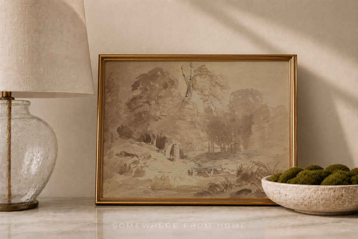 A framed sepia-toned landscape painting with a gold frame, styled on a marble surface next to a glass lamp and a bowl of green moss.