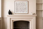 A framed abstract art print with textured white and gray brushstrokes hangs on a beige wall above a cream-colored fireplace mantel decorated with black vases and a small white bowl.