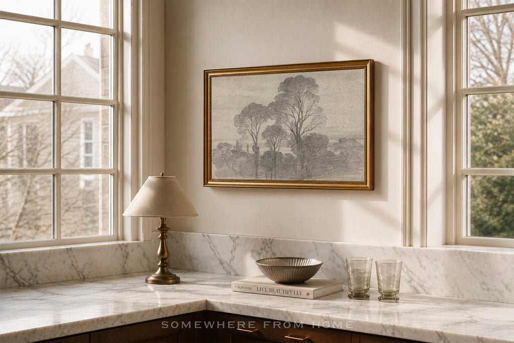A framed landscape art print with trees hangs on a beige wall above a marble kitchen counter with a lamp, bowl, and glasses in a sunlit room.