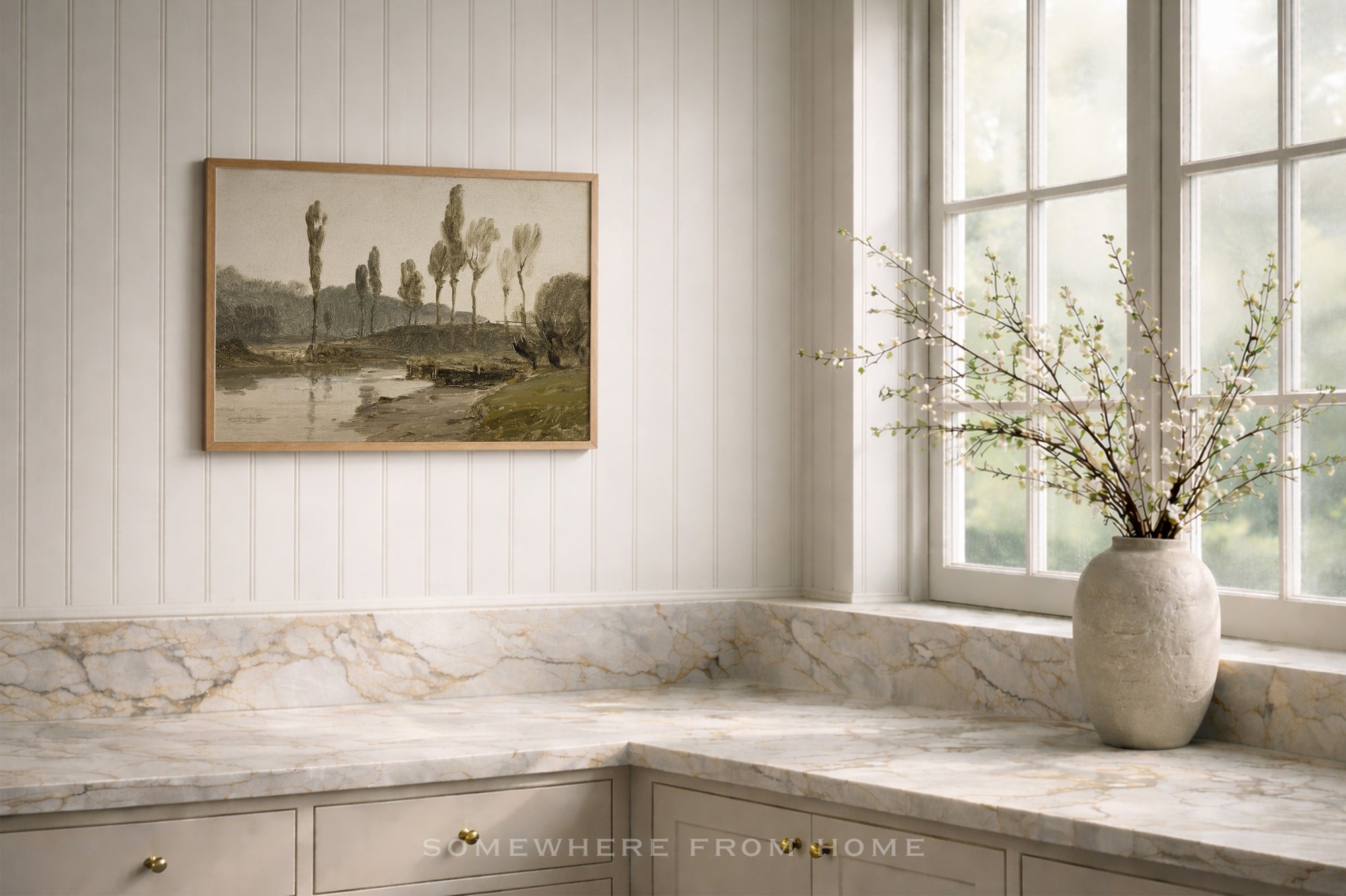 A framed landscape art print hangs on a white paneled wall above a marble countertop in a bright kitchen. A large textured vase with delicate branches sits by the window.