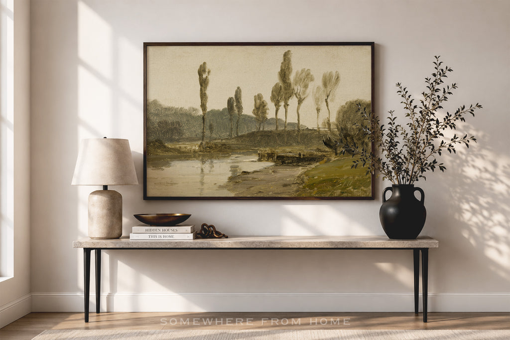 A framed landscape art print hangs on a beige wall above a console table in a styled room. The print depicts a soft, muted countryside scene with trees, a river, and distant hills in beige, green, and brown tones.