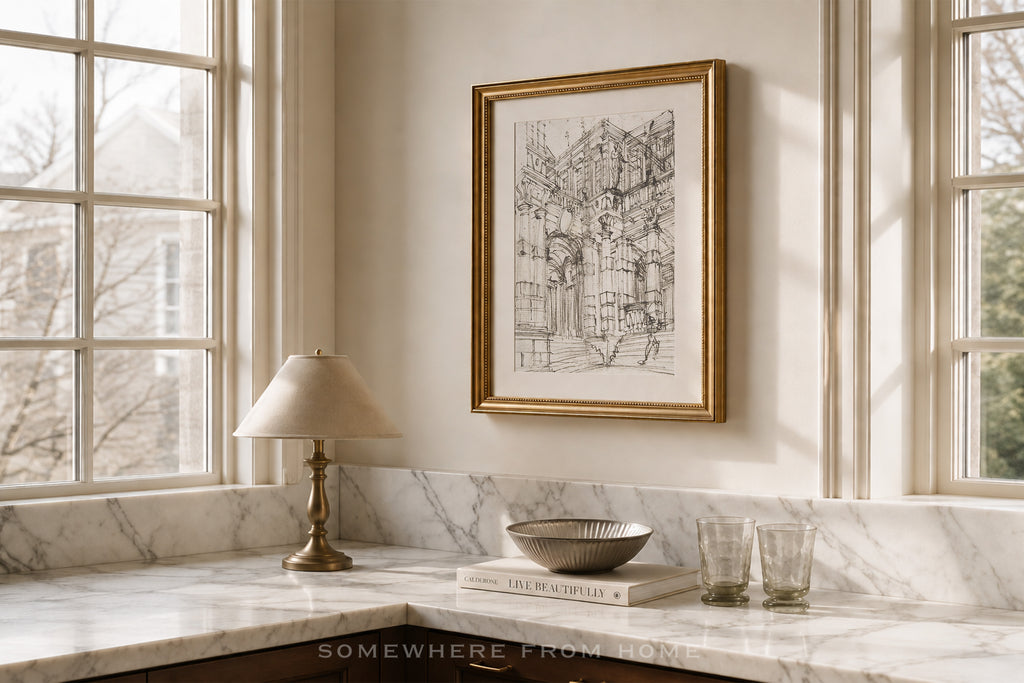 A framed black and white architectural sketch print hangs on a cream wall above a marble kitchen counter with a lamp, bowl, and glasses.