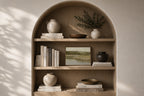 An arched wooden built-in shelf with three tiers, styled with ceramic vases, books, a small bowl, and a framed landscape print against a warm beige wall with soft leaf shadows.