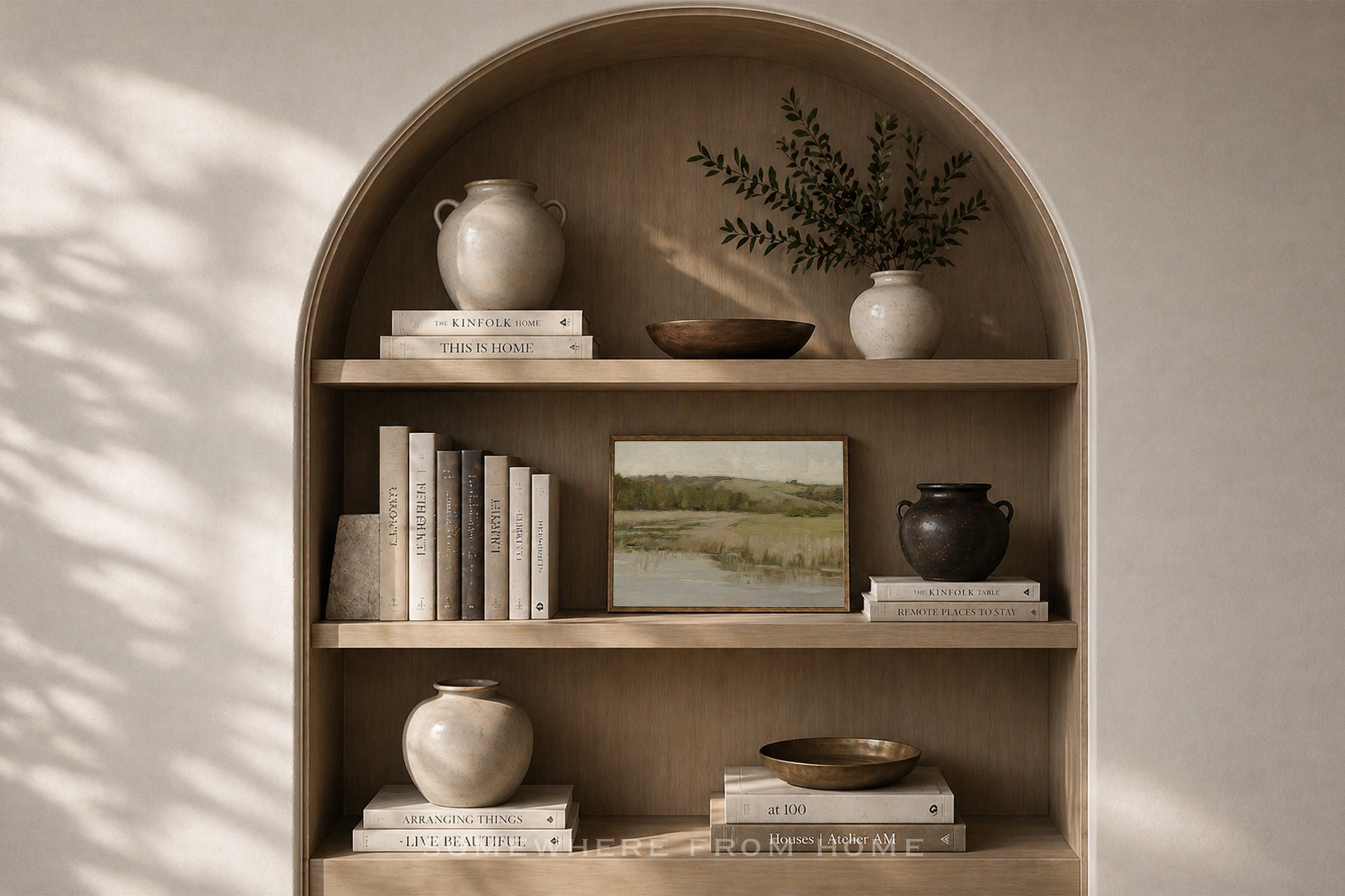 An arched wooden built-in shelf with three tiers, styled with ceramic vases, books, a small bowl, and a framed landscape print against a warm beige wall with soft leaf shadows.