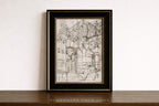 A framed black and white architectural sketch print leaning against a white wall on a dark wooden surface. The print features a detailed drawing of an ornate building facade, enclosed in a black frame with a gold inner border.