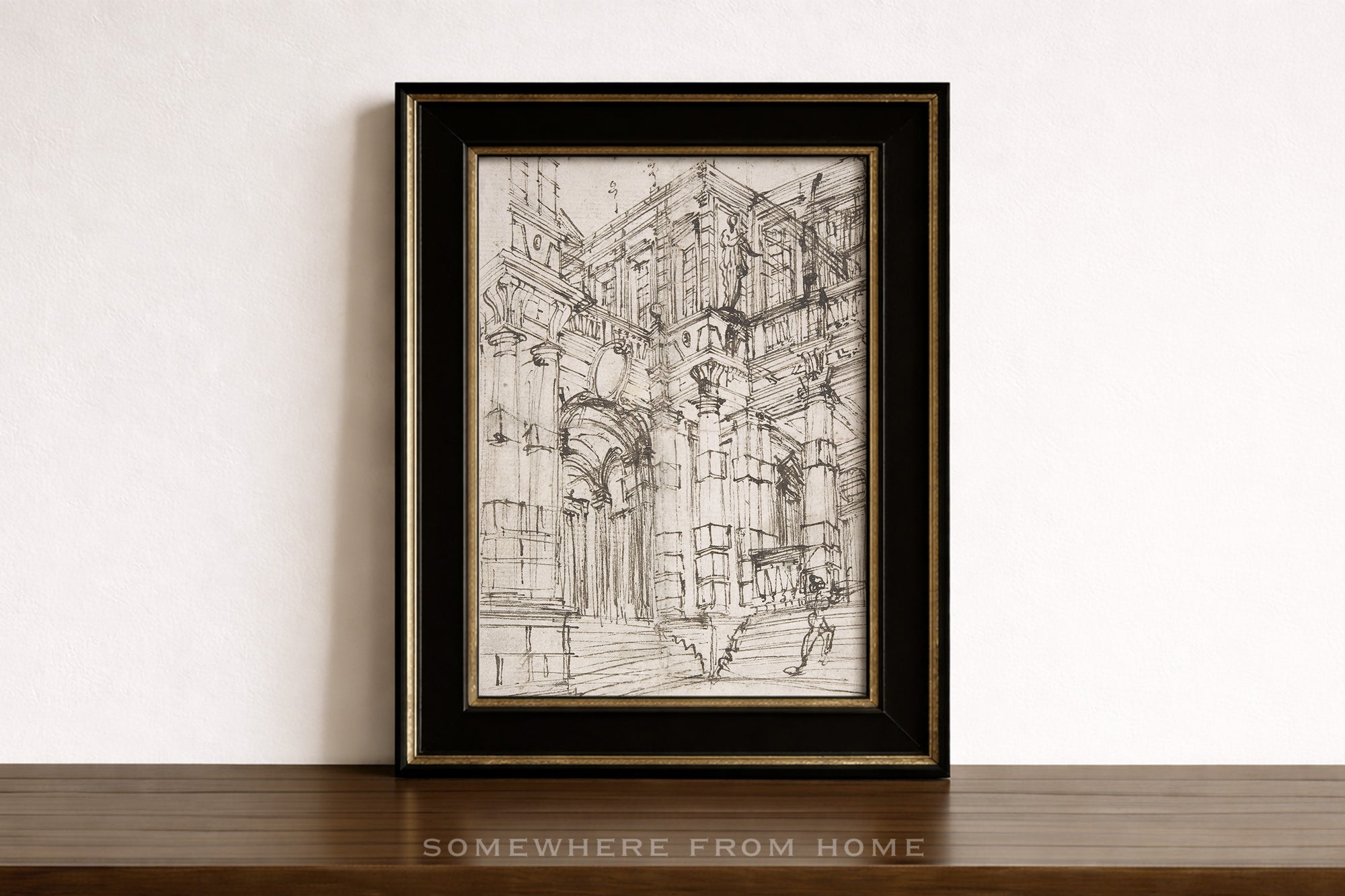 A framed black and white architectural sketch print leaning against a white wall on a dark wooden surface. The print features a detailed drawing of an ornate building facade, enclosed in a black frame with a gold inner border.