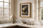 A framed still life art print with a white flower and dark green leaves, set in a gold frame and hanging on a cream wall above a marble kitchen counter with a lamp and books.