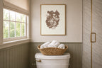 A framed botanical art print in brown and cream hangs on a beige wall above a white toilet in a styled bathroom, with a woven basket of white towels on top. The print appears to be the product being showcased in a real interior setting.