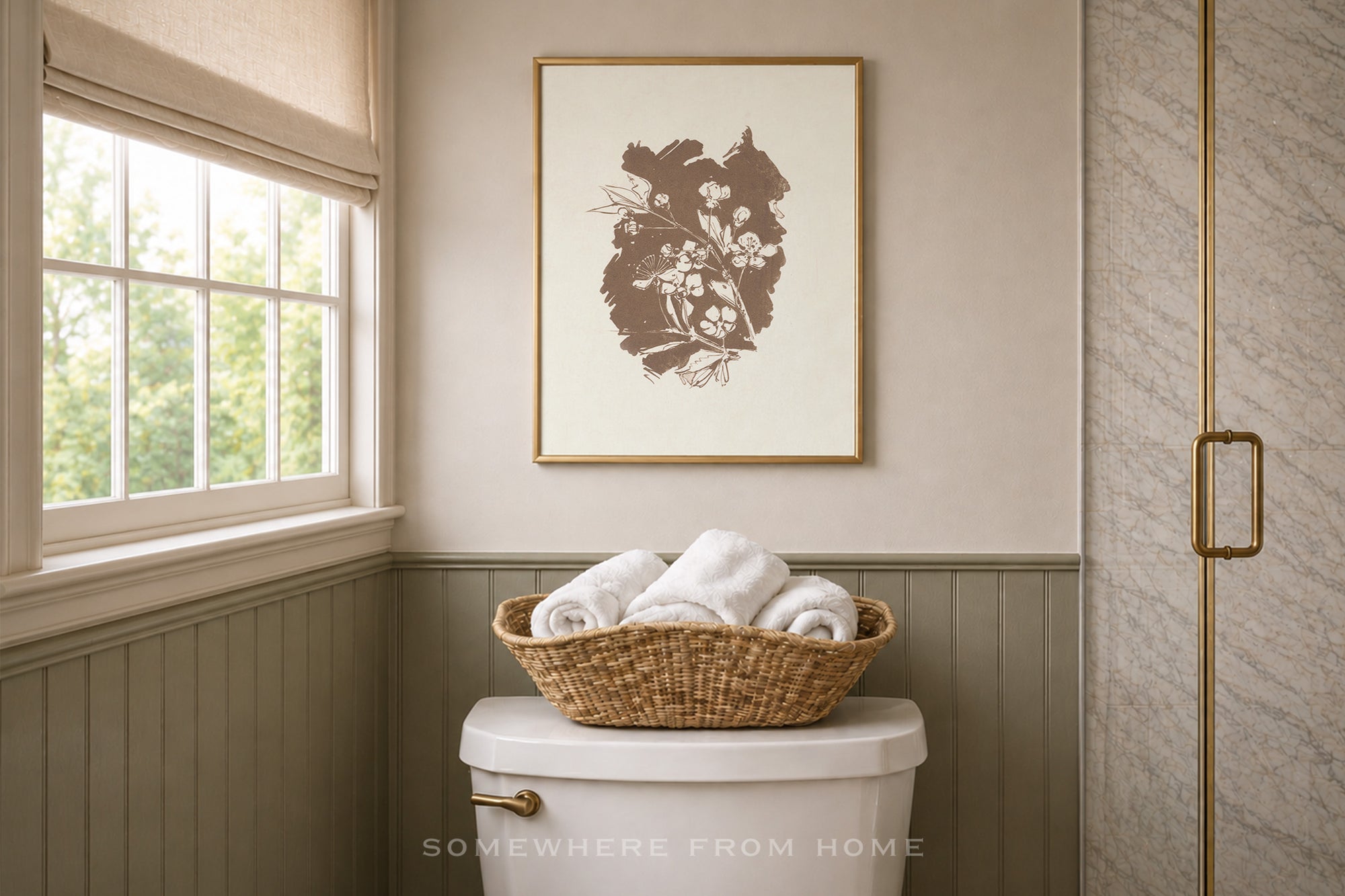 A framed botanical art print in brown and cream hangs on a beige wall above a white toilet in a styled bathroom, with a woven basket of white towels on top. The print appears to be the product being showcased in a real interior setting.