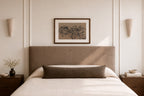 A styled bedroom scene featuring a bed with a tall upholstered headboard, white bedding, a dark brown lumbar pillow, a framed botanical print on the wall, and matching wall sconces.