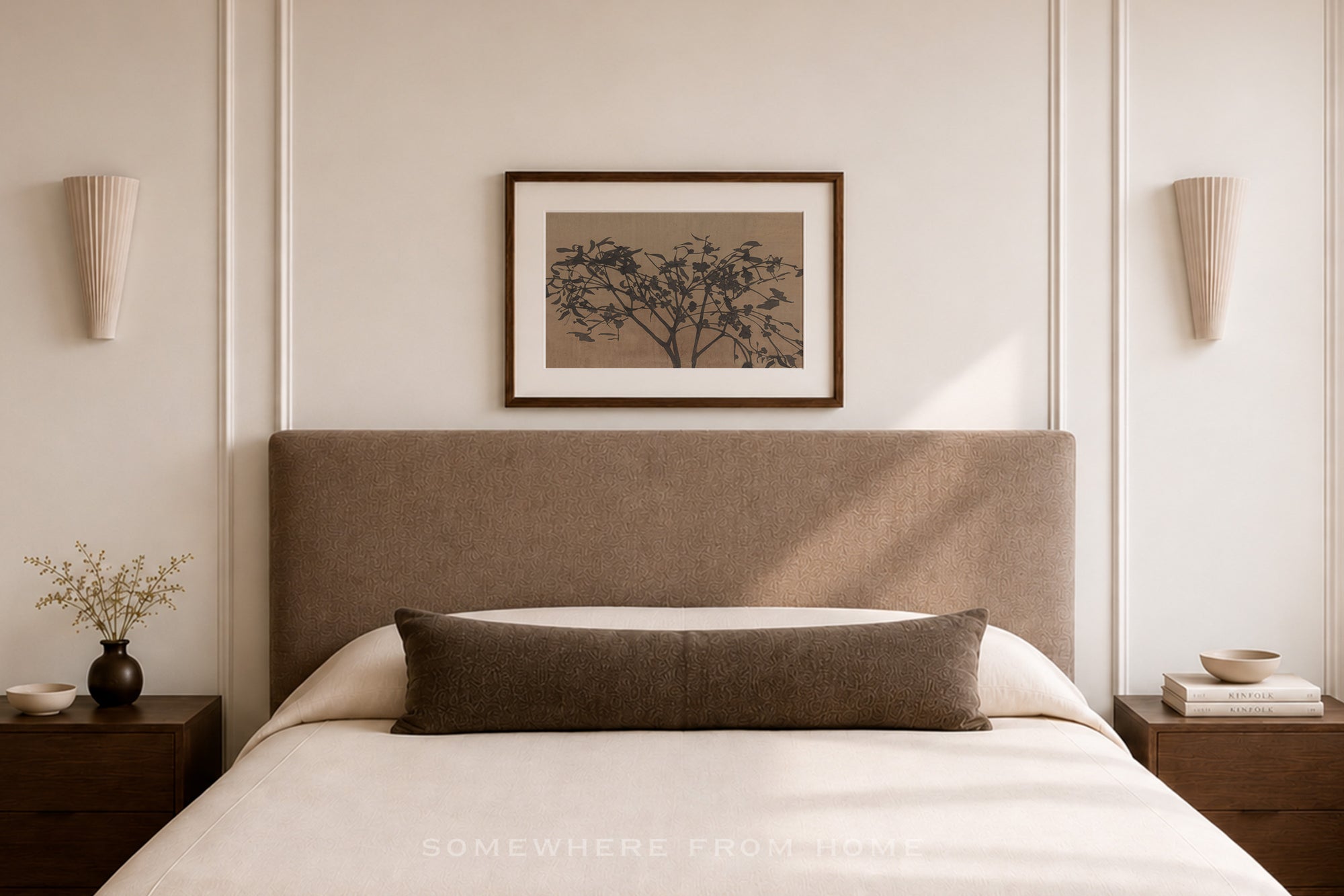 A styled bedroom scene featuring a bed with a tall upholstered headboard, white bedding, a dark brown lumbar pillow, a framed botanical print on the wall, and matching wall sconces.