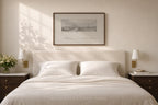 A styled bedroom scene featuring a neatly made bed with white linens and pillows, flanked by matching dark wood nightstands with wall-mounted lamps. Above the bed hangs a framed monochrome landscape print.