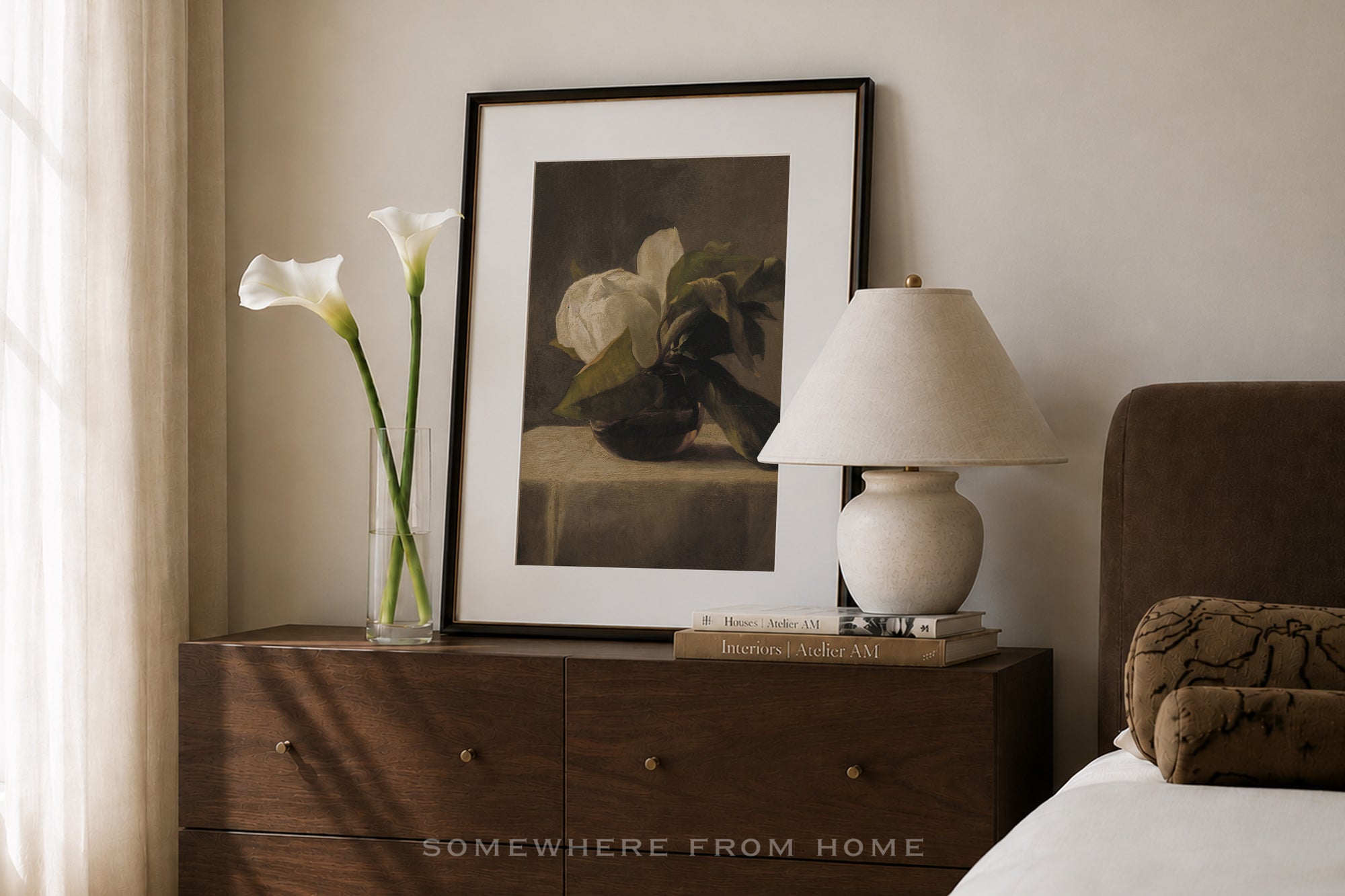 A framed still life art print featuring white lilies, styled on a dark wood nightstand next to a table lamp and a vase with calla lilies in a bedroom setting.